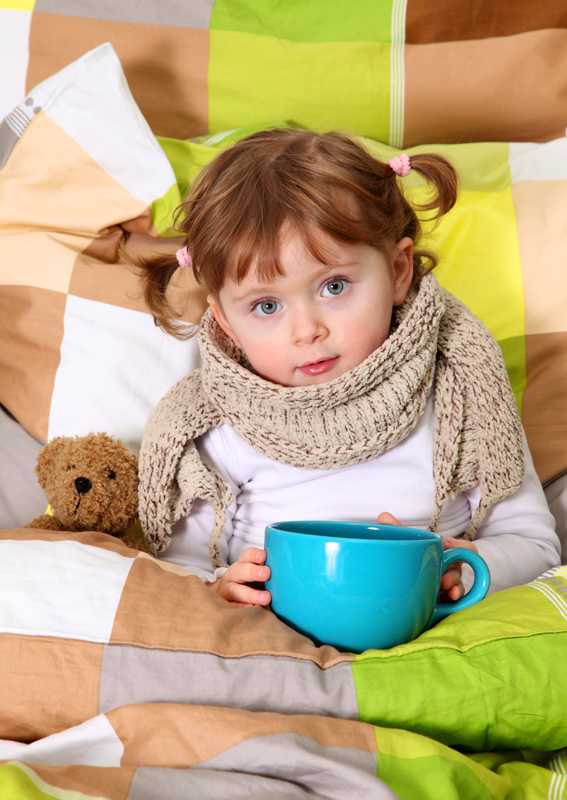  Image d'une petite fille malade entourée de couette dans un lit. Elle tient un bol bleu dans ses mains et garde son doudou à gauche de l'image. 