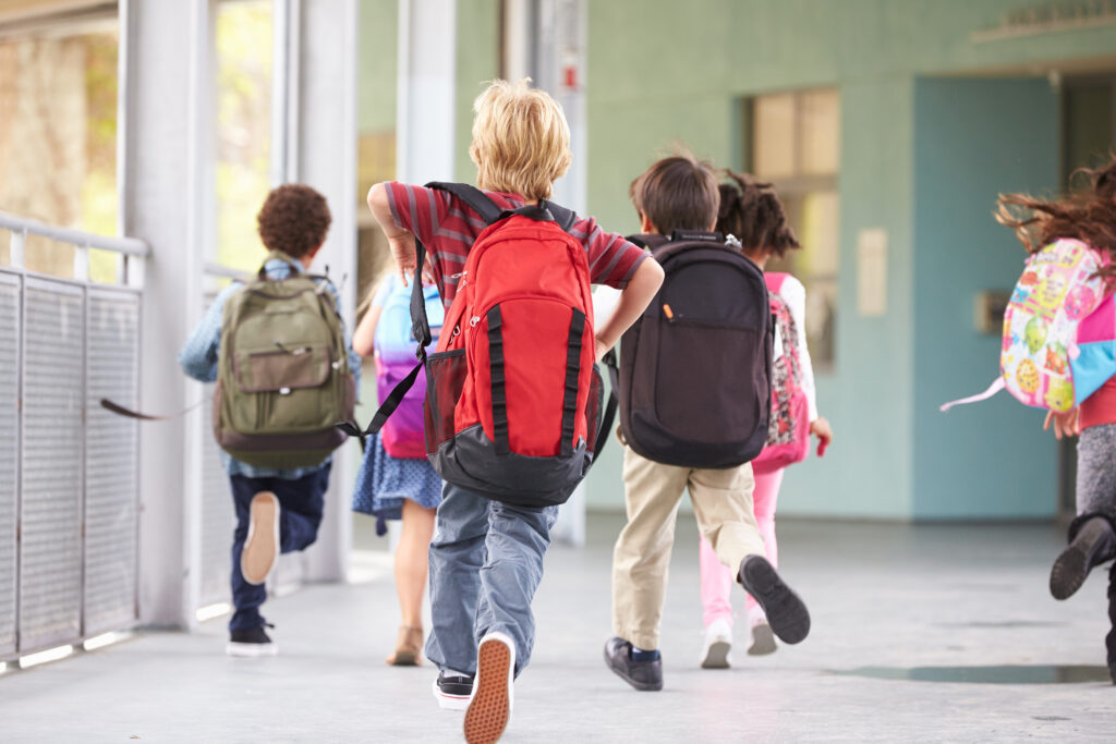 Image où l'on voit des enfants qui courent vers la sortie de l'école en fin de journée.
On les voit tous de dos avec de grands sac à dos sur leurs épaules.